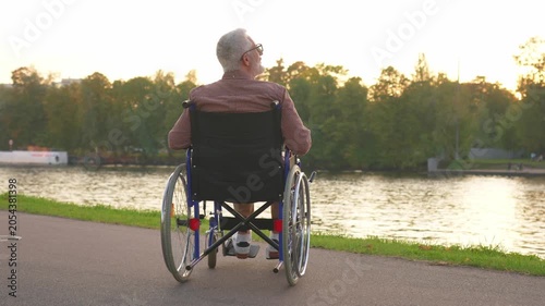 Sad senior man sitting in wheelchair walking on road in city park outdoor, back view. Old paralyzed man in chair for people with disability feel depressed. Lonely elderly sick man sits on wheelchair