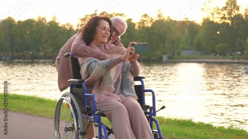 Senior couple woman in wheelchair caregiver old man making selfie enjoying walking. Elderly family man supporting embracing woman in chair for people with disability taking photo. Rehabilitation