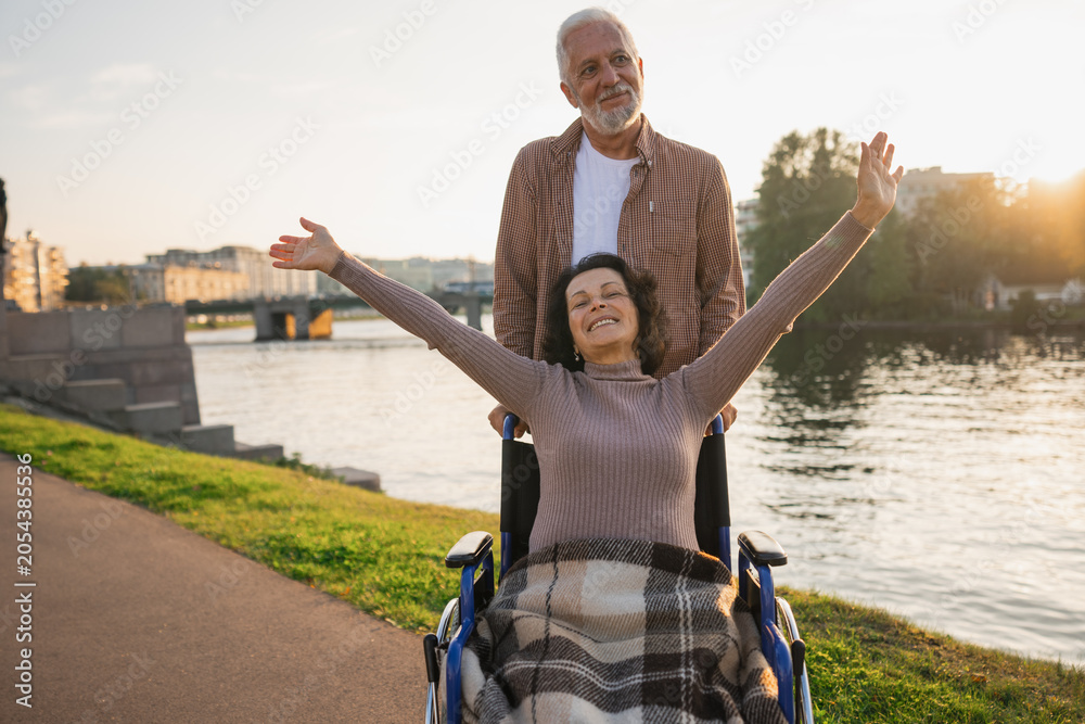 Fototapeta premium Senior woman in wheelchair walking with caregiver old man on road. Family couple man husband supporting taking care of paralyzed woman wife in chair for people with disability outdoor. Rehabilitation