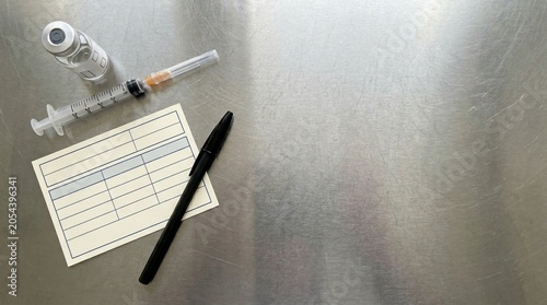 Medical supplies including a syringe with needle, vial and a record card with a pen on a metallic surface