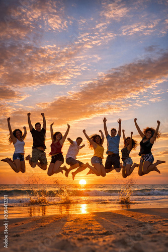A group of people joyfully jumping in the air with arms raised, celebrating together at sunset on a beach with waves and clouds in the background.