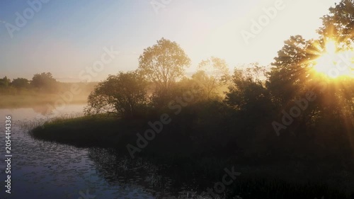Sunbeams shining through trees silhouettes on a misty riverbank at sunrise