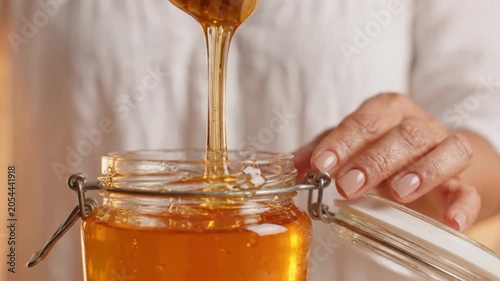 A person carefully scooping honey from a glass jar with a wooden honey dipper in a bright and clean environment.
