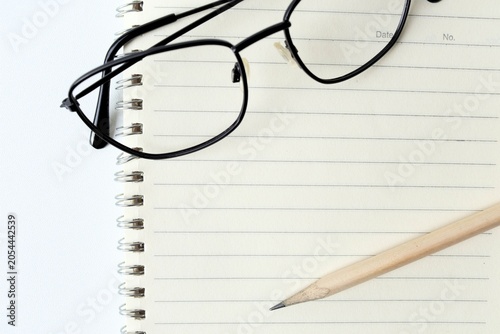 Top view of black frame eyeglasses and a wooden pencil on an open blank lined spiral notebook, isolated on white background, concept of education, studying, and workspace planning.