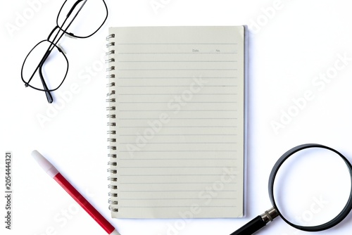 Top view of eyeglasses, magnifying glass, and red marker arranged around an open blank lined spiral notebook creating a frame with copy space, isolated on white background.