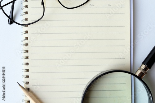 ​Top view of eyeglasses, a wooden pencil, and a magnifying glass arranged around an open blank lined spiral notebook, creating a frame with copy space, isolated on white background.