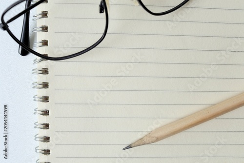 Top view of black frame eyeglasses and a wooden pencil on an open blank lined spiral notebook, isolated on white background, concept of education, studying, and workspace planning.
