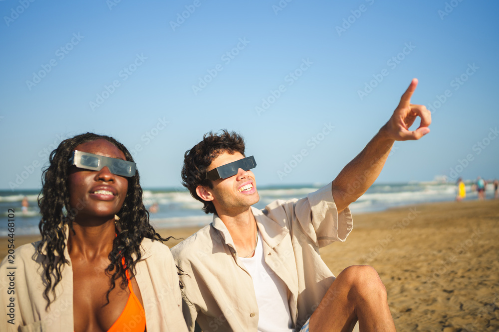 Naklejka premium Two people on beach wearing solar eclipse glasses, man pointing to sky