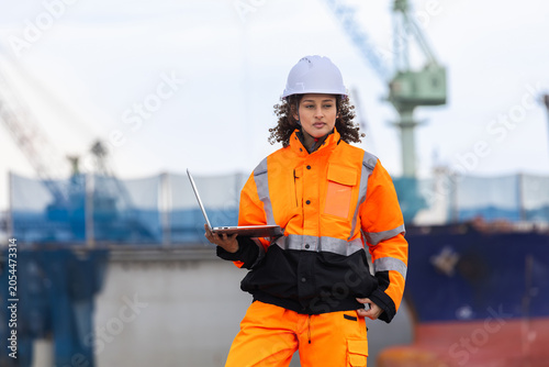 Professional female engineer holding laptop monitoring shipping operations with digital equipment at harbor, Portrait of a female site supervisor in high visibility uniform working at commercial port