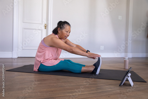 Senior Asian woman stretching forward on exercise mat at home, wearing pink top, following tablet