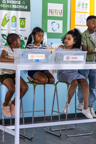Diverse children sorting plastic and glass bins at white table in classroom with recycling posters