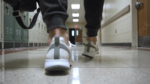Individual wearing gray sweatpants and white sneakers walks down a school hallway, carrying a black bag, with lockers lining the walls on either side