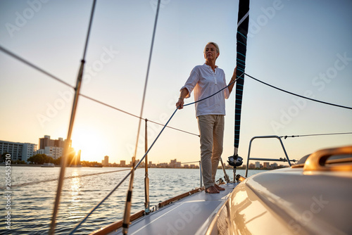 A woman stands confidently on a sailboat, adjusting the sail while the sun sets over a bustling city skyline. The warm light reflects on the water. Generative AI.
