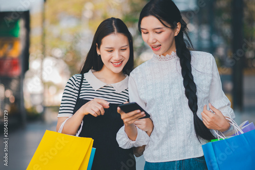 Two women are looking at a cell phone while holding shopping bags