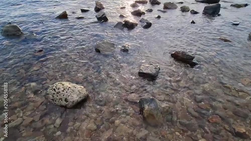 Scenic wide shot of various sized rocky boulders and pebbles scattered throughout clear shallow sea water during daytime.
