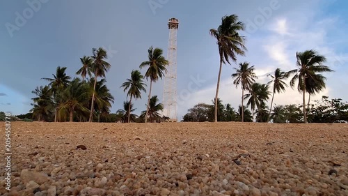 A low-angle shot of a tall white steel skeleton lighthouse tower surrounded by tropical coconut palm trees on a coarse pebble sand beach.