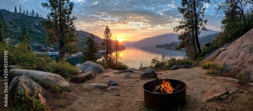 Lakeside campfire at sunrise with mountains and trees framing tranquil water
