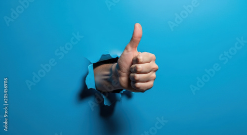 Thumbs Up Symbol Rising From Hole in Bright Blue Wall for Positive Endorsement Approval and Positive Confirmation