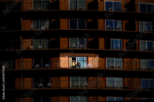 Bild auf Leinwand A lone individual stands on a sunlit balcony against a backdrop of dimly lit units