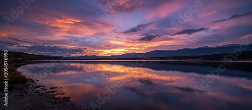 Vivid sky ablaze with sunset colors reflects on a calm lake, mountains in distance