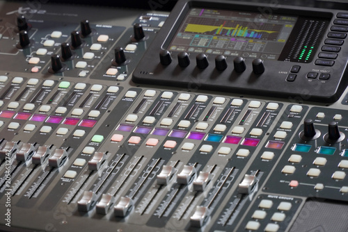Foto Close-up of a professional digital audio mixing console with illuminated colorful LED buttons, knobs, and channel faders in a dark music recording studio or broadcasting control room