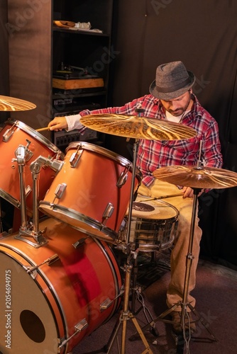 Fotografie Mid-adult Hispanic musician with hat practicing beats on a drum set indoorsMid-a