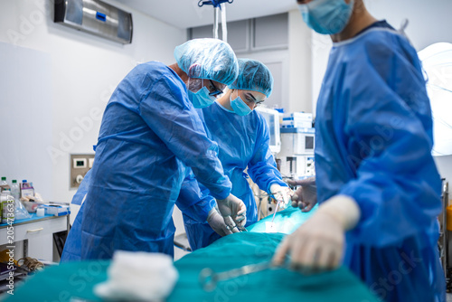 Team of focused medical doctors and surgeons performing critical operation in hospital operating room. Professional surgical group in blue protective uniforms and masks working on patient care.