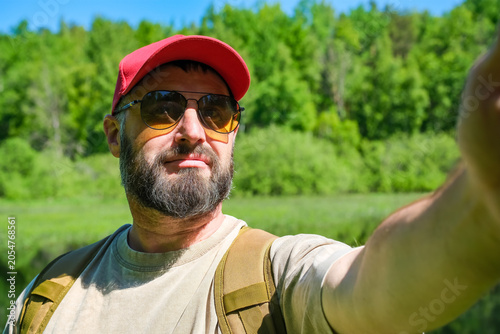 Foto Adult bearded man in sunglasses takes outdoor selfie near green riverside, summer travel, self portrait and social media activity