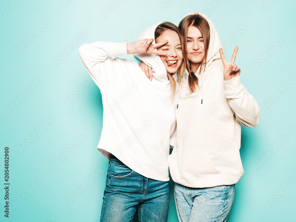 Fototapeta premium Two young beautiful brunette smiling hipster in trendy summer clothes. Sexy carefree women posing near blue wall. Trendy and positive models having fun in sunglasses