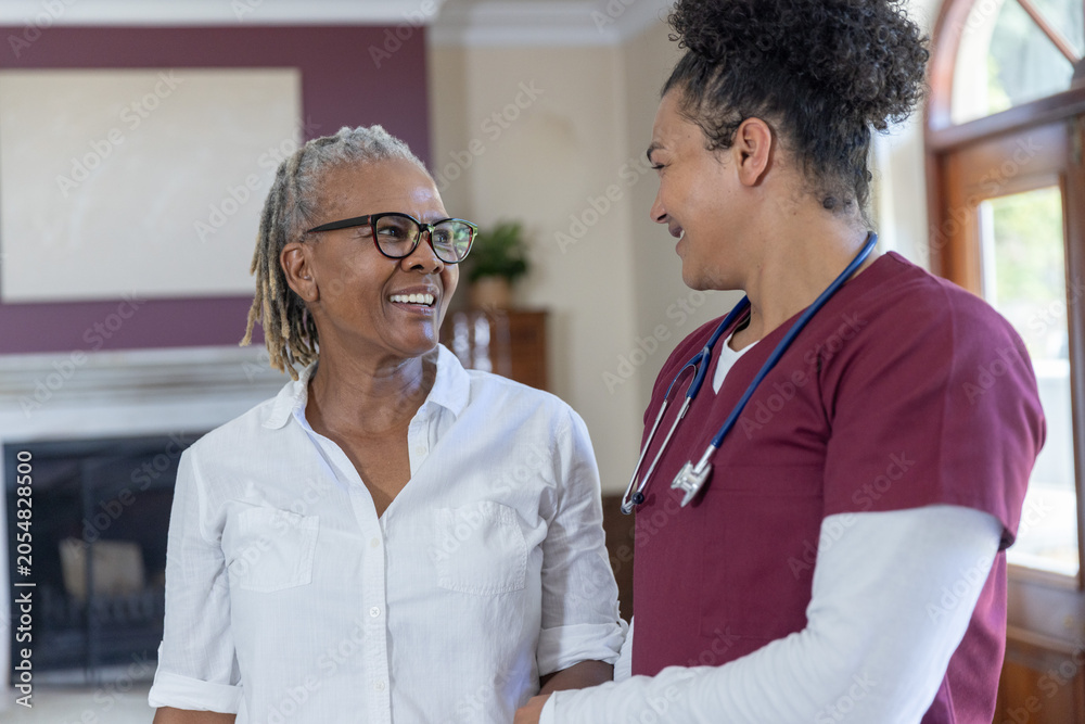 Obraz premium Senior African American woman and caregiver in scrubs standing, talking in living room, stethoscope