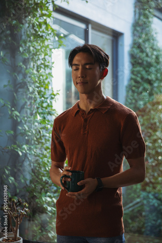 Adult Asian man standing in narrow courtyard holding dark green mug, rust-brown polo, potted plants