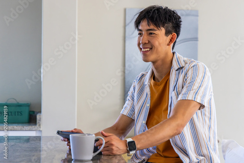 Asian man sitting at kitchen counter smiling, holding phone and mug, showing smartwatch, copy space