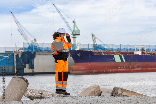 Female engineer with laptop at shipyard terminal, Logistics coordinator monitoring shipping vessel operations at a commercial harbor