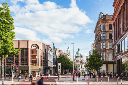A bustling day shot captures pedestrians walking down Donegall Place in Belfast city center, looking past historic and modern storefronts toward the green dome of Belfast City Hall.