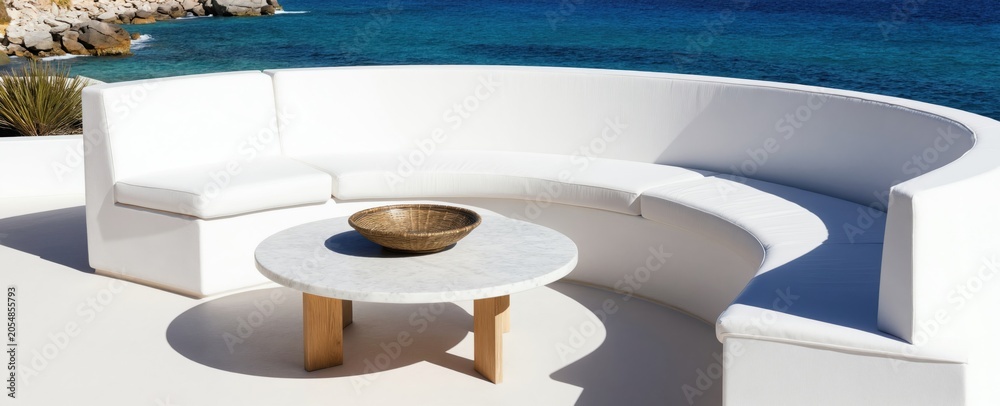 Naklejka premium Modern white curved sofa and marble table on sunny terrace with ocean view. Outdoor lounge area offers tranquil relaxation and coastal escape with blue sea and rocky shore.