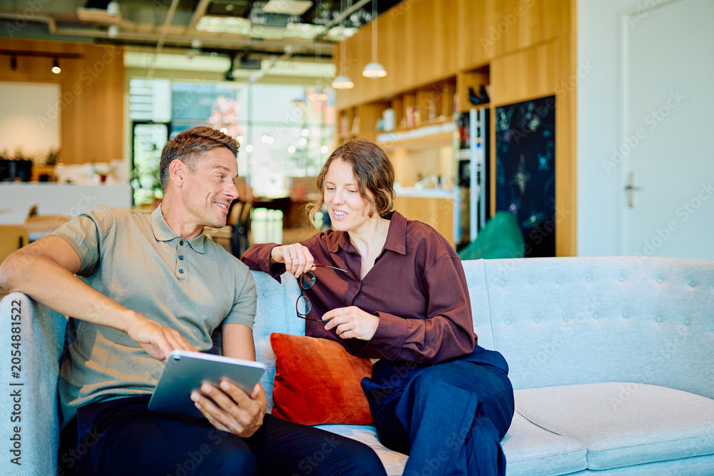 Naklejka premium Two business colleagues sitting on a couch, discussing work and sharing ideas while using a digital tablet in a casual office setting