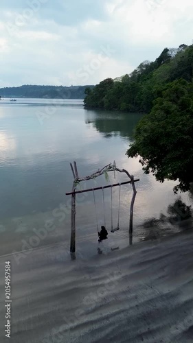 Moslem asian woman person enjoys a relaxing moment on a wooden swing at the beach in Weh island Sabang Indonesia, overlooking the ocean and the horizon on a sunny day.  Travel at the resort
