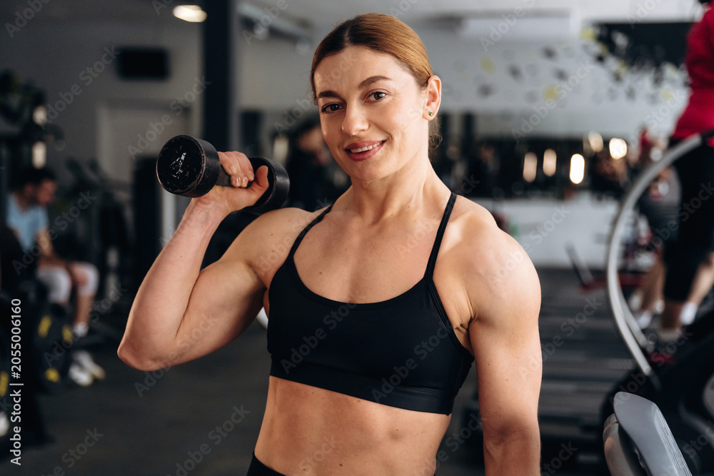 Fototapeta premium Female fitness enthusiast with muscular build holds dumbbell in gym, workout equipment and people exercising visible in the background
