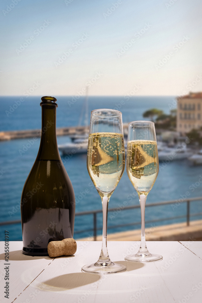 Fototapeta premium Champagne flutes and bottle on balcony overlooking seaside marina. Concept of luxury travel, celebration toast, romantic riviera getaway, premium sparkling wine and coastal resort leisure.