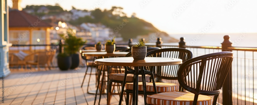 Fototapeta premium Coastal town view from a charming bistro terrace featuring elegant seating and sunlit ocean scenery.