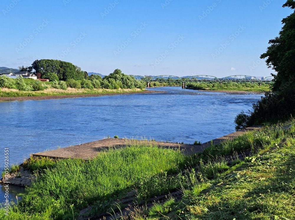 Fototapeta premium 岩手県花巻市・青空と新緑に包まれた北上川の「イギリス海岸」と遠くに望むトラス橋の風景
