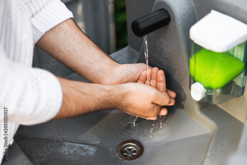 Fotografie Close-up of a man washing his hands under running water at an outdoor portable sink with a green liquid soap dispenser, focusing on hygiene and sanitation