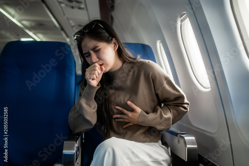 Young woman coughing and holding her chest while sitting by the window on an airplane, showing travel illness, discomfort, and in-flight health concern during a flight.