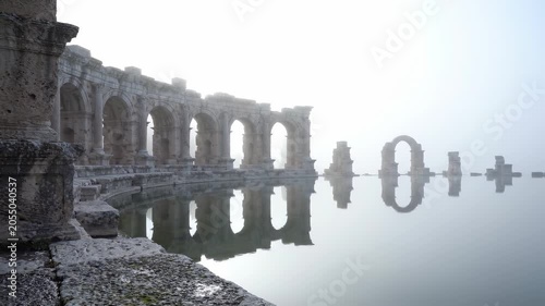 Misty dawn blankets the serene waters, casting ethereal reflections of ancient ruins. The delicate interplay of light and shadow evokes a sense of timeless beauty and mystery.