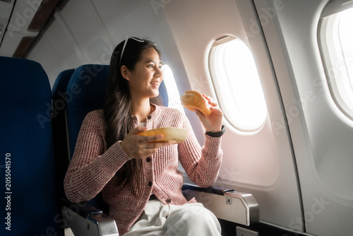 Young woman sitting by the airplane window enjoying a snack during a flight, holding bread and a plate in a bright cabin seat, showing comfort, travel, and relaxed in-flight dining.