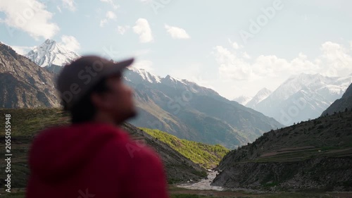 4K shot of a defocused hiker wearing hat looking at the Himalayan mountain valley with snowy mountain peaks and river flowing in front of it at Miyar Valley Trek in Lahaul, Himachal Pradesh, India.