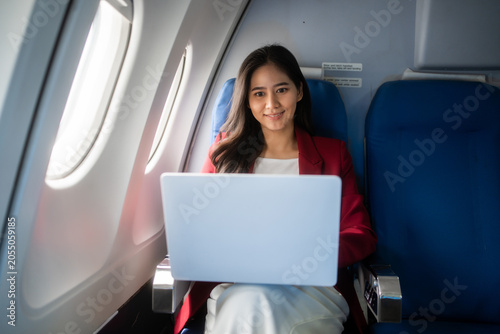 Smiling young woman using a laptop while seated by the window on an airplane, showing business travel, remote work, comfort, and modern mobility.