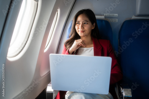 Smiling young woman using a laptop while seated by the window on an airplane, showing business travel, remote work, comfort, and modern mobility.