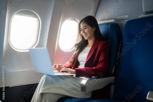 Smiling young woman using a laptop while seated by the window on an airplane, showing business travel, remote work, comfort, and modern mobility.