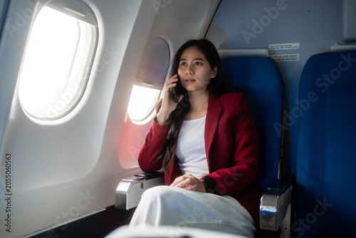 Young woman in a red blazer sits by an airplane window, talking on her phone during a flight. calm travel scene with natural light, business trip, communication, and modern air travel.